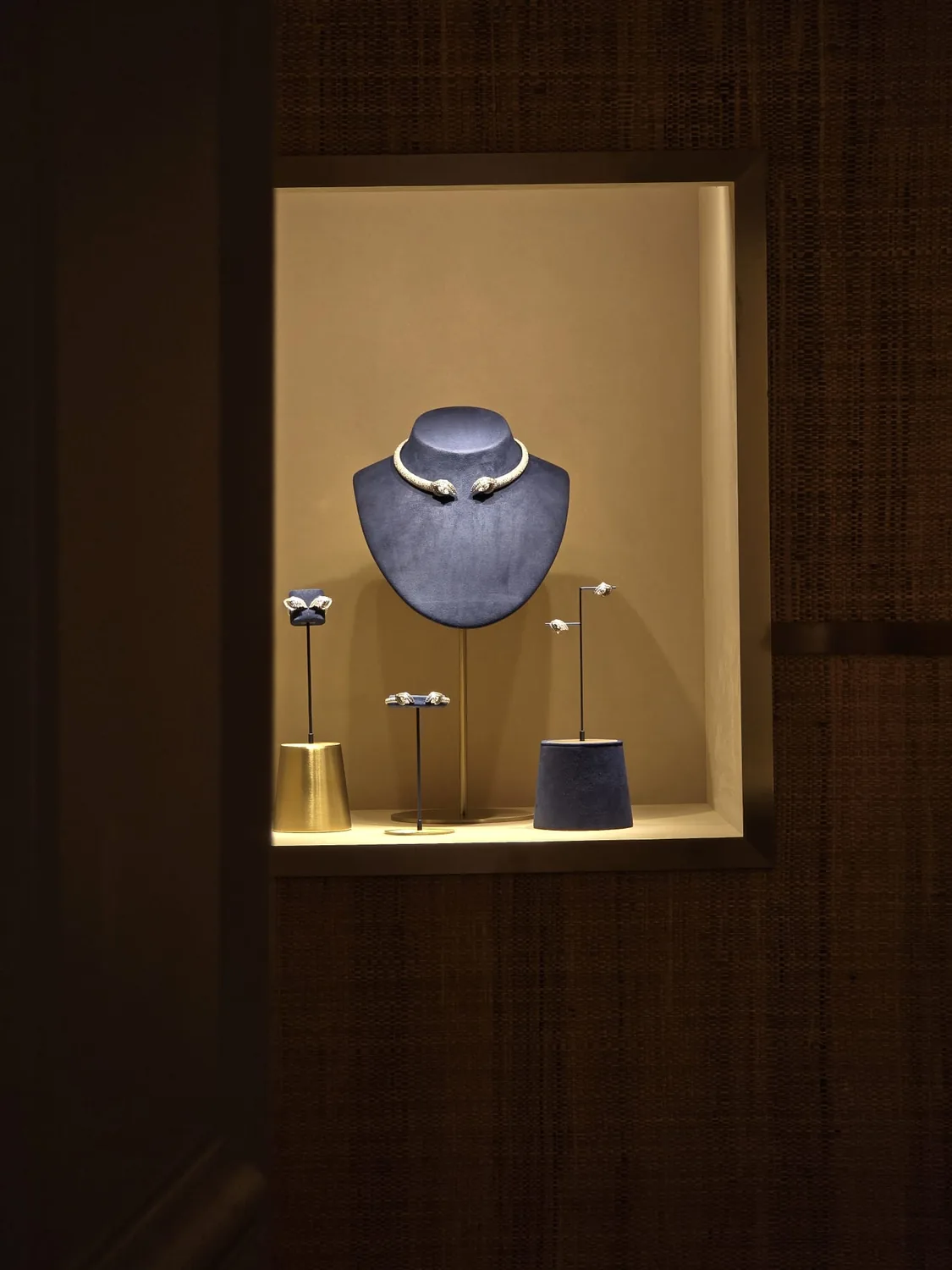 A diamond necklace on a navy bust in a lit boutique niche, with earrings on stands beside it.